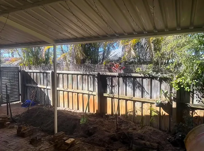 Backyard under a patio before, with a weathered timber fence and a dug-out garden bed